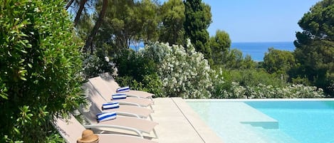 A poolside area with lounge chairs and a table, surrounded by greenery and overlooking the sea.