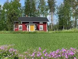Una casa rossa con una porta gialla, immersa in un prato verde e circondata da fiori viola.