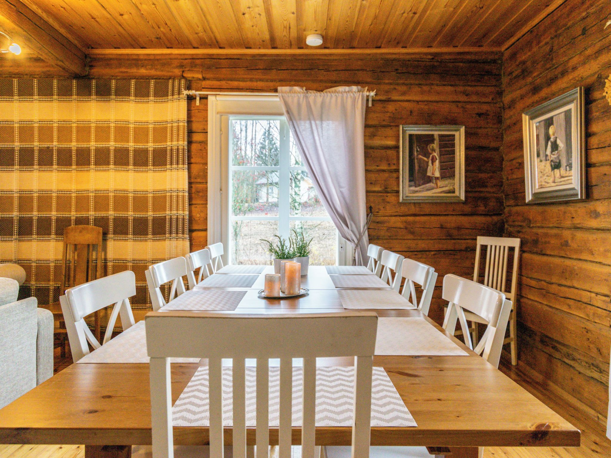 A wooden cabin interior with a dining table set for six, white chairs, a window with curtains, and framed pictures on the walls.