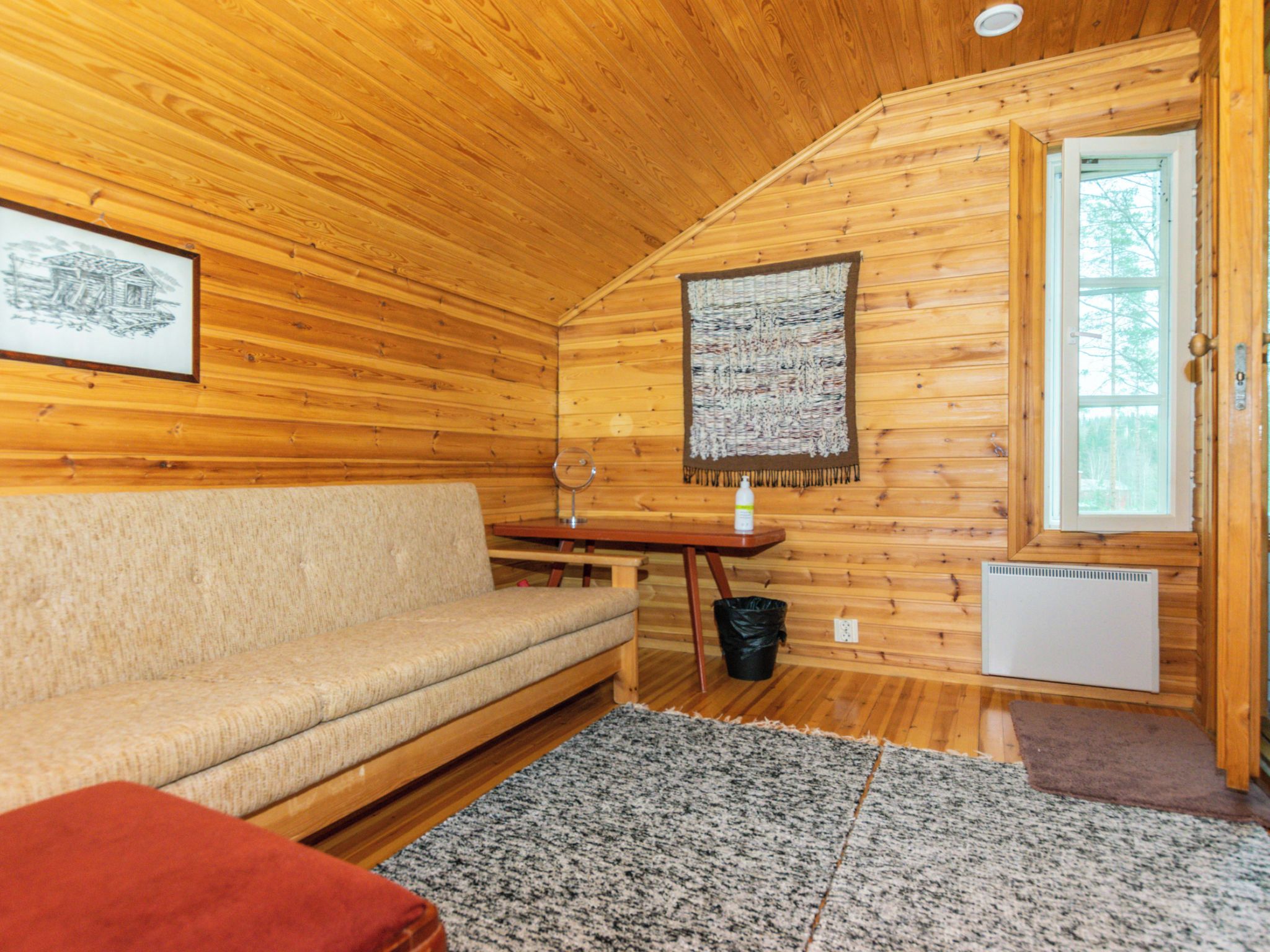 A cozy cabin interior with a wooden ceiling, a sofa, a table, and a window.