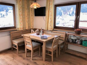 Un coin repas chaleureux avec une table et un banc en bois, un téléviseur fixé au mur et une fenêtre donnant sur un paysage enneigé.