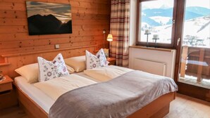 Ein Schlafzimmer mit Holzwänden, einem Bett, floralen Kissen, Bergblick und einem fenster mit Vorhängen.