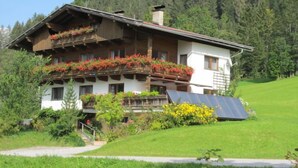 Eine zweistöckige Chalet mit Holzbalkonen, die mit roten Blumen geschmückt sind, umgeben von üppiger Vegetation und einem bewaldeten Hügelhang.