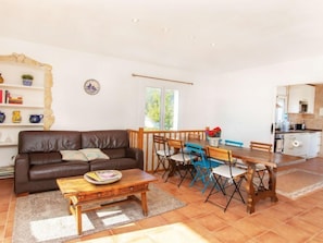 A cozy living room with a brown leather sofa, a wooden coffee table, and a dining area with a wooden table and blue chairs.