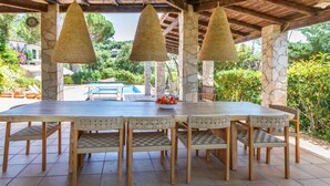 A covered outdoor dining area with a stone pillar, a pool in the background, and a view of greenery. - El Suro by Interhome (Calonge)