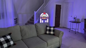 A living room with a grey sofa, a checkered pillow, a jukebox, and a small table with a lamp. - THUNDERDOME (awesome Dome House) Unique home with Beautiful views/Comfy and Cozy (Erie)