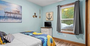 A bedroom with a bed, a television on a dresser, a painting of a pier, and a window with curtains.