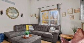 A living room with a grey sofa, a wooden coffee table with a plant, a window with curtains, and framed pictures on the wall.
