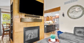A living room with a fireplace, a television mounted on the wall, a dining area with chairs, and a kitchen in the background. - Red Timbers Near Beaches and Wineries (Traverse City)