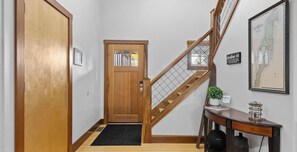 A hallway with a wooden door, a staircase with a wooden railing, a welcome sign, a framed map, and a desk with a plant.