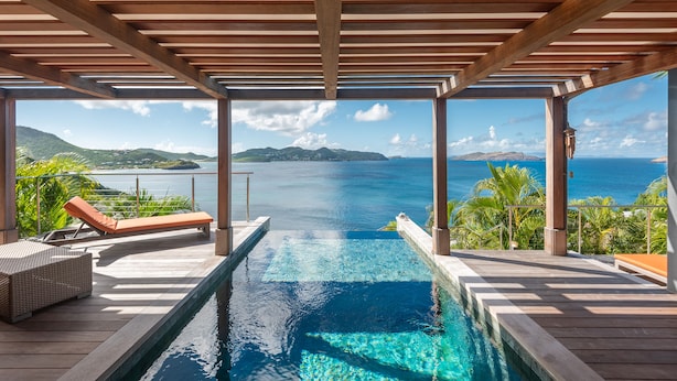 Une piscine à débordement avec une terrasse en bois, des chaises longues et une vue sur l’océan et les montagnes.