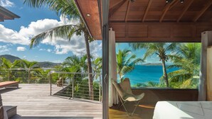 A balcony with a wooden deck, a cushioned chair, and a view of the ocean and palm trees. - Villa Bayugita | Ocean View - Located in  Exquisite Pointe Milou with Private Pool (Pointe Milou)