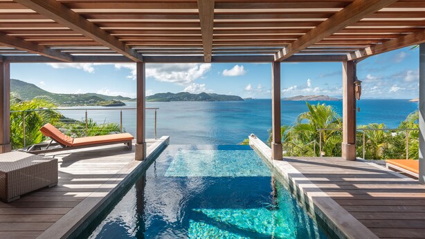 An infinity pool with a wooden deck, lounge chairs, and a view of the ocean and mountains. - Villa Bayugita | Ocean View - Located in  Exquisite Pointe Milou with Private Pool (Pointe Milou)