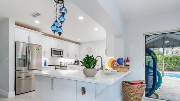A modern kitchen with white cabinetry, stainless steel appliances, a central island, and a decorative blue chandelier. - Portal2TheSea Only STEPS to the BEACH (Pompano Beach)