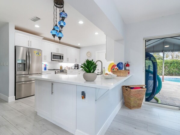 A modern kitchen with white cabinetry, stainless steel appliances, a central island, and a decorative blue chandelier. - Portal2TheSea Only STEPS to the BEACH (Pompano Beach)
