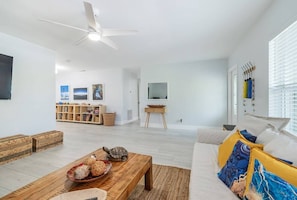 Amplia sala de estar con ventilador de techo, un sofá con cojines coloridos, una mesa de centro de madera con objetos decorativos, y una estantería llena de libros y un televisor sobre un soporte.