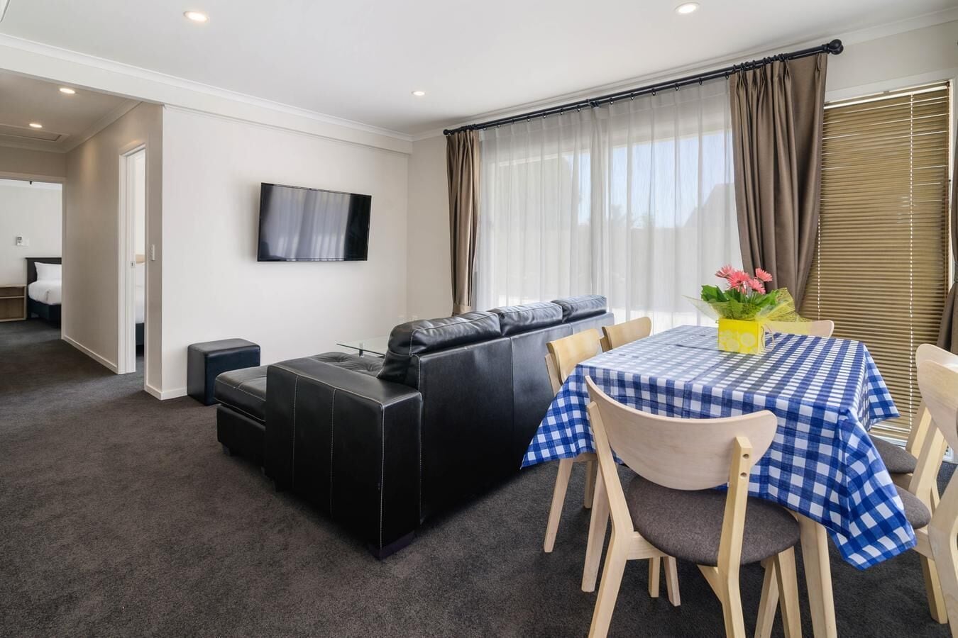 Un salon moderne avec un canapé en cuir noir, un téléviseur à écran plat fixé au mur et un coin repas avec une nappe à carreaux.
