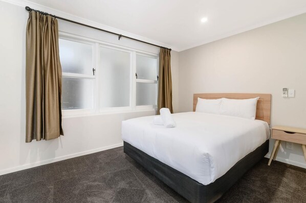 Una habitación de hotel con una cama grande, una mesita de noche, una ventana con cortinas y una pared lisa.