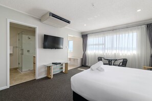 Une chambre d’hôtel avec un grand lit, un téléviseur fixé au mur, une douche en verre, une petite table avec des chaises et une fenêtre avec des rideaux.