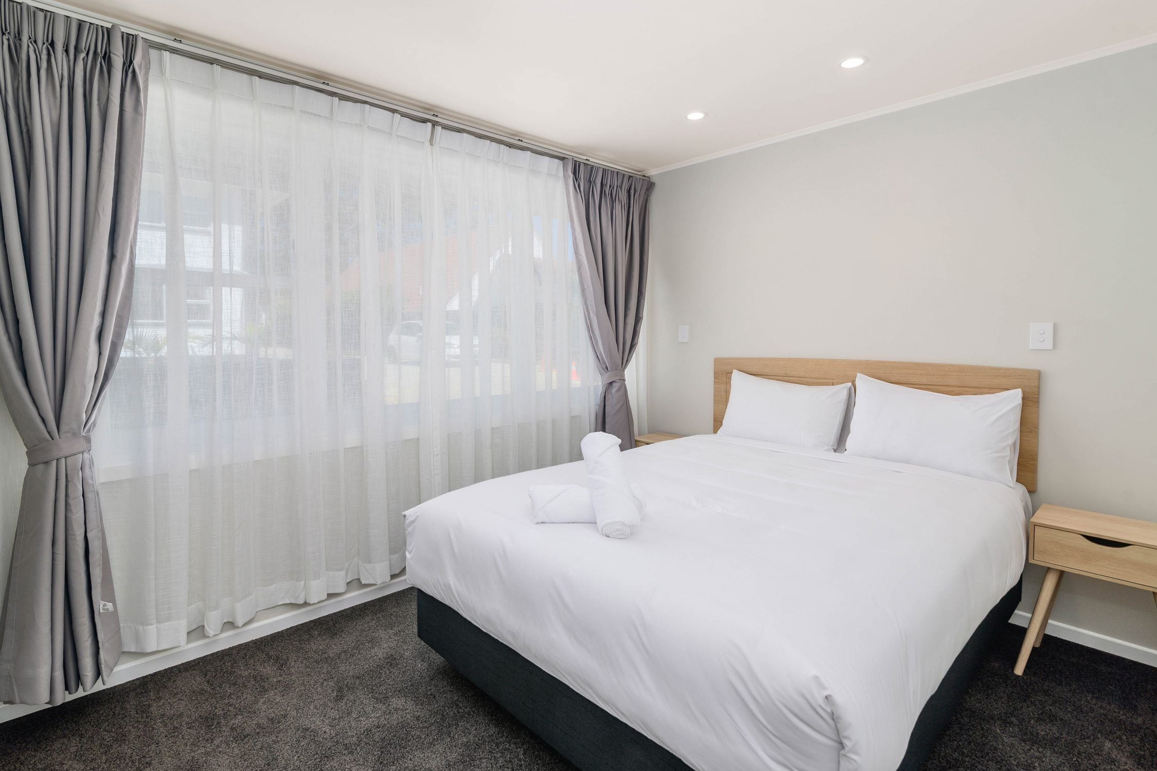 Una habitación de hotel con una cama grande, cortinas grises, un cabecero de madera y una mesita de noche de madera.