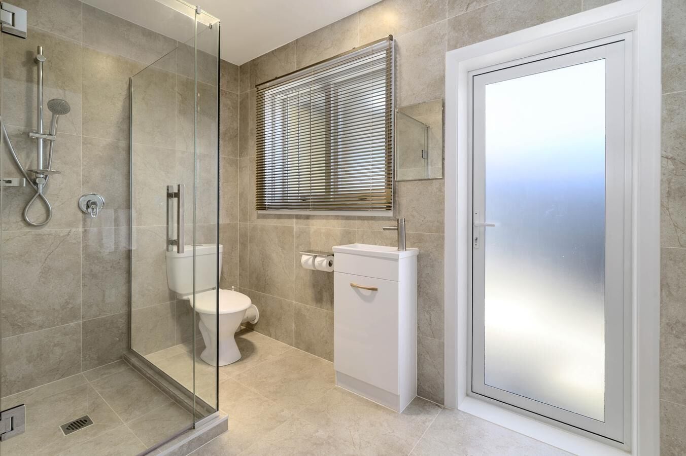 Un baño moderno con ducha envuelta en vidrio, inodoro, lavamanos y puerta de vidrio satinado.