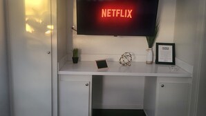 Un escritorio blanco con un televisor montado que muestra el logotipo de Netflix, una pequeña planta en maceta, un objeto decorativo y un certificado enmarcado.