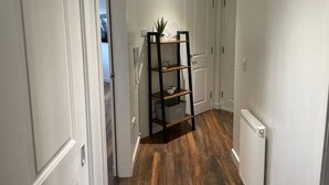 Un couloir aux murs blancs, doté d’un plancher en bois et d’une étagère en métal noir sur laquelle sont posés divers objets.
