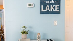A room with a light blue wall, a white console table with a plant, a jar, and binoculars, and a sign that reads "at the LAKE". - Private Lakefront House with Hot Tub, Kayaks and Beach (Selkirk)