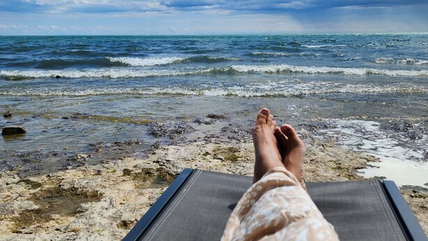 A beach view with a lounge chair and a person's legs. - Private Lakefront House with Hot Tub, Kayaks and Beach (Selkirk)