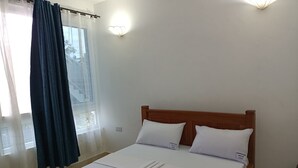 Un lit bien fait, avec du linge de lit blanc et des oreillers, une tête de lit en bois et une fenêtre ornée de rideaux légers.