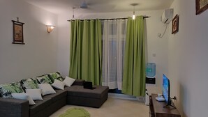 Un salon comprenant un canapé d’angle, un distributeur d’eau, un ventilateur de plafond et un téléviseur posé sur un meuble en bois.