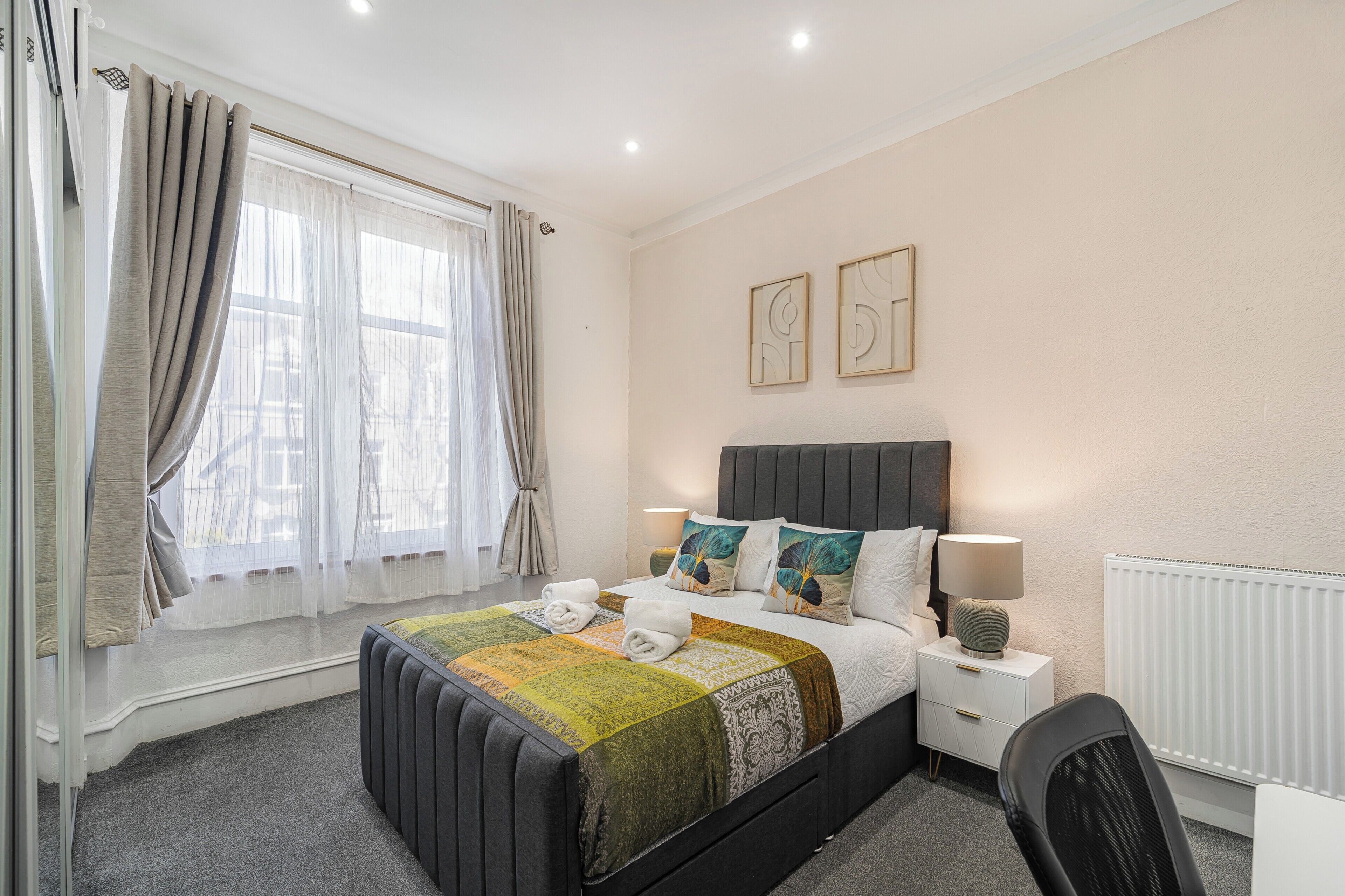 Un dormitorio con cama, mesitas de noche, una silla, ventana con cortinas y decoración en la pared.
