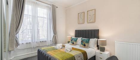 Un dormitorio con cama, mesitas de noche, una silla, ventana con cortinas y decoración en la pared.