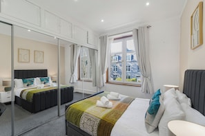 A modern hotel room with a large bed, a mirror, a bedside table, a window with curtains, and a city view. - Comfortable West End Flat with Parking Easy Access (Aberdeen)