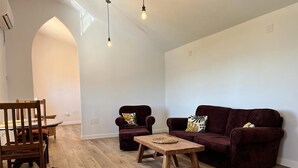 Ein Wohnzimmer mit Holzboden, einem dunkelroten Sofa, zwei Sesseln, einem Holztisch und einem Esstisch mit Stühlen.