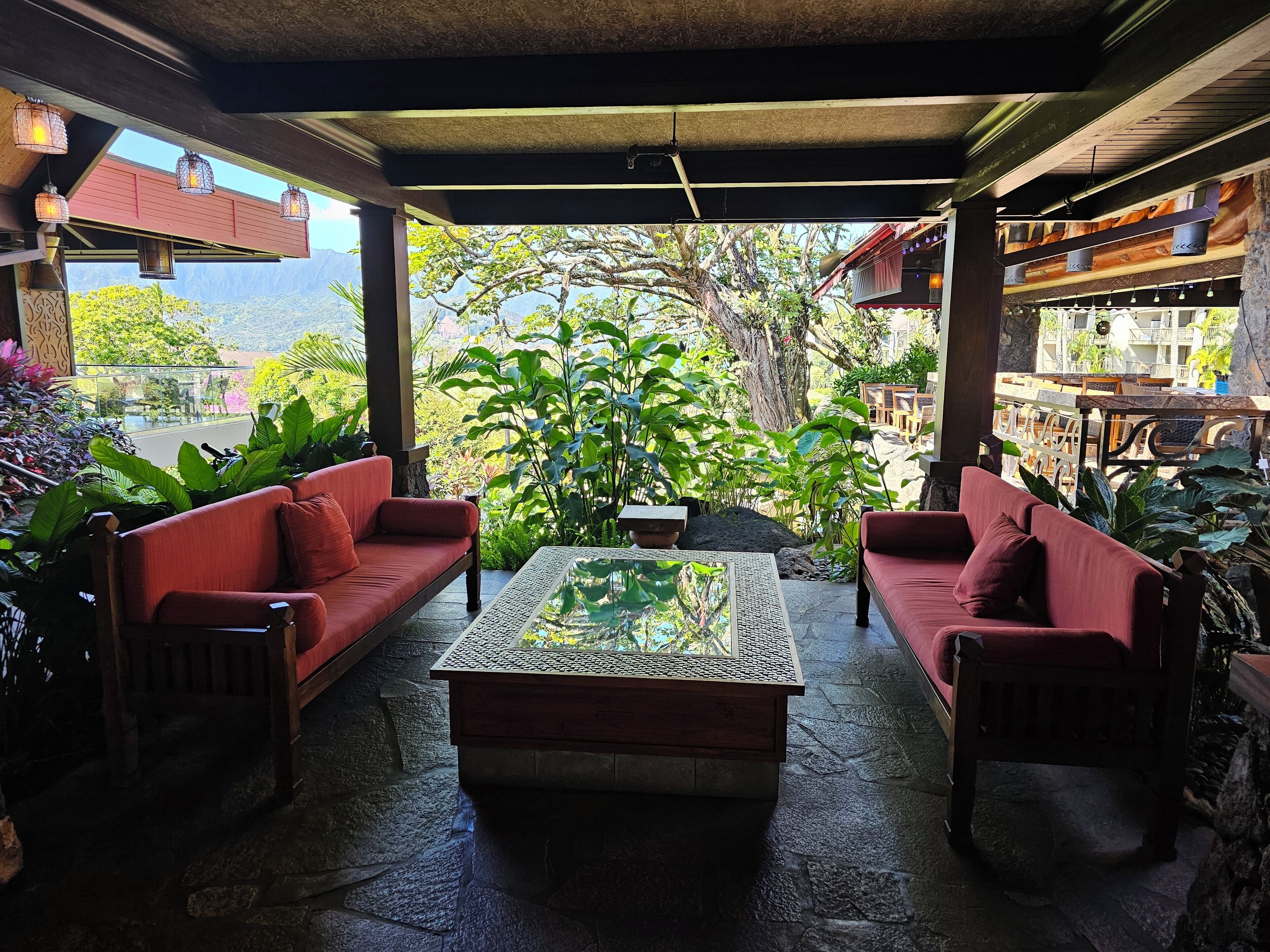 Un espace extérieur aménagé, abrité et doté de canapés rouges et d’une table basse en verre, entouré d’une végétation luxuriante et offrant une vue sur une chaîne de montagnes.
