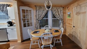 Ein Zimmer mit Holzwandverkleidung, einem runden Esstisch und weißen Stühlen, einer kleinen Küchenzeile inklusive Geschirrspüler sowie einem Fenster mit Vorhängen.
