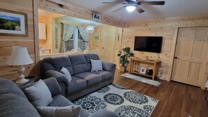 Ein gemütliches Wohnzimmer mit einer grauen Couch, einem kleinen Tisch mit einer Lampe, einem Fernseher und Holzwänden.