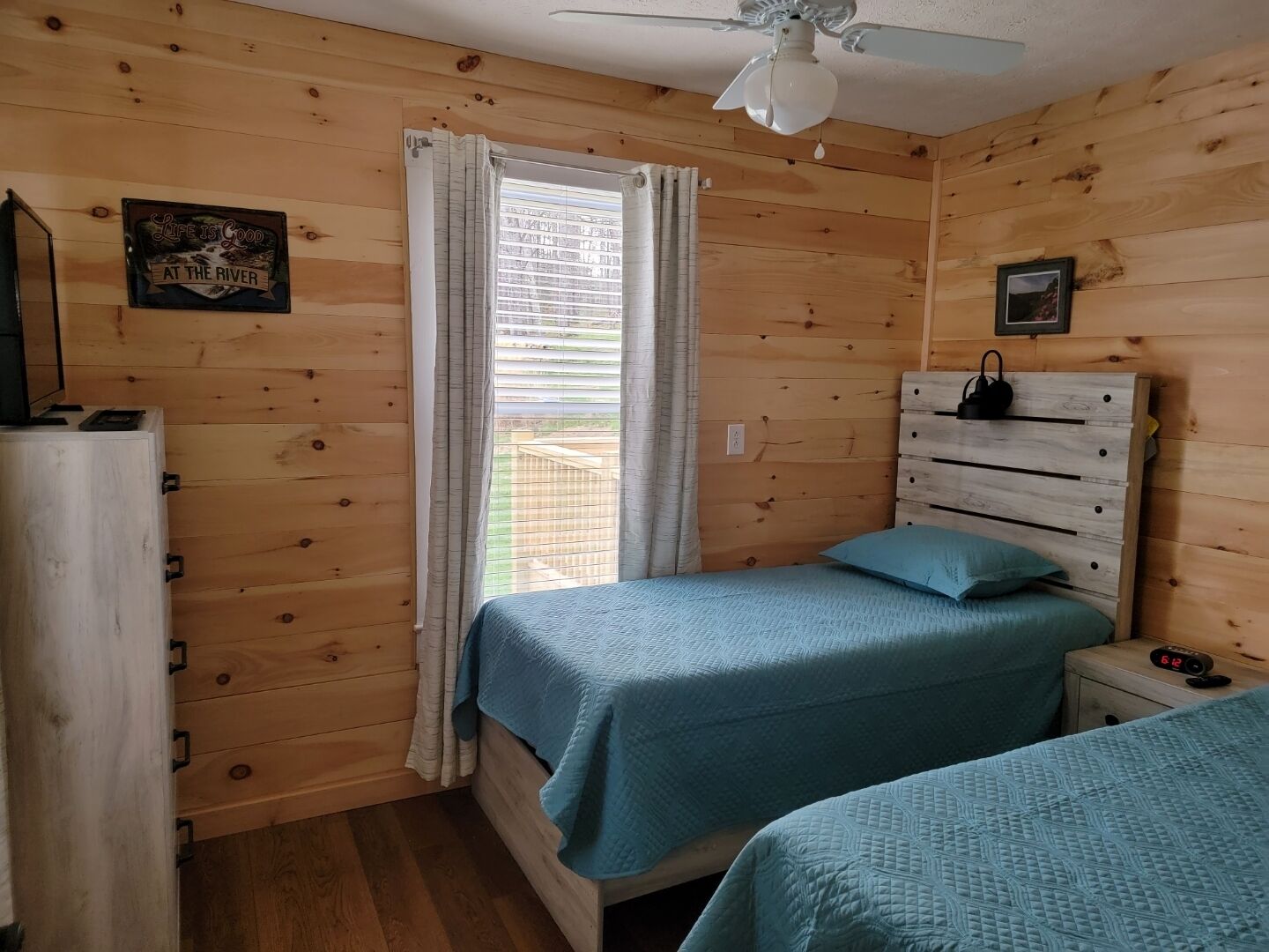 A room with wooden walls and a ceiling fan. There is a bed with a blue bedspread and a nightstand with a clock. A window with curtains is visible, and a sign on the wall reads "Life is Good at the River."
