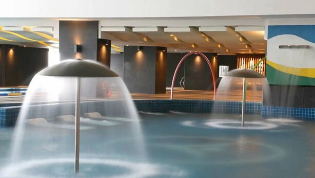 Un espace piscine intérieur moderne doté de plusieurs éléments aquatiques, notamment des fontaines et un toboggan aquatique central.