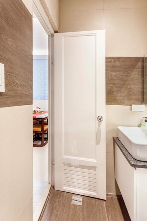Een smalle gang met een houten vloer, een witte deur en een badkamer met een wastafel en spiegel.