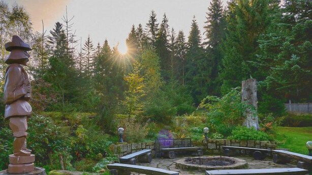Un foyer en pierre, entouré de bancs, se trouve au milieu d’un jardin arboré.