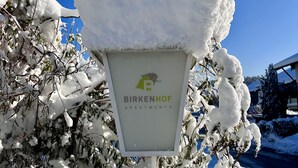 Een bordje voor Appartementen Birkenhof, met een met sneeuw bedekt dak en takken.