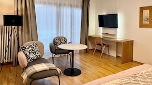 Une chambre d’hôtel moderne équipée d’un canapé, de deux fauteuils, d’une petite table ronde, d’un bureau en bois, d’un téléviseur à écran plat et d’un radiateur.