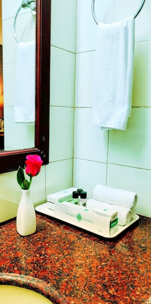 A bathroom with a white towel hanging on a metal ring, a white vase with a rose, and a tray with toiletries including a box labeled "Kovilakom Residency." - Kovilakam Residency Private Limited (Kozhikode)