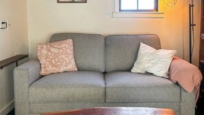 A grey sofa with floral and plain pillows, a wooden coffee table with a remote control, and a window with a light fixture.