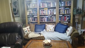 A cozy living room with a leather armchair, a sofa with patterned cushions, and a wooden coffee table with a decorative item. A bookshelf filled with books is in the background.