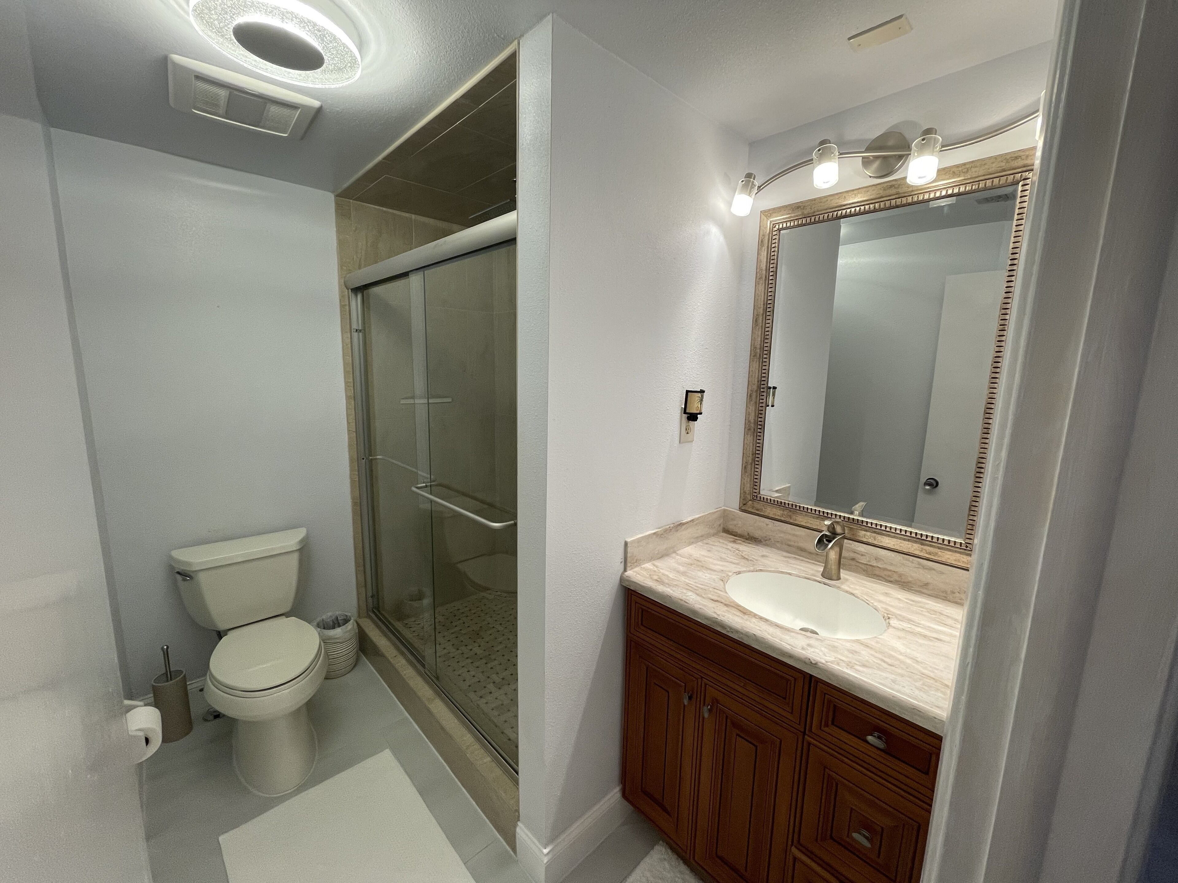 Une salle de bain moderne avec une douche à parois de verre, des toilettes blanches et un meuble-lavabo doté d’un grand miroir et d’un luminaire.