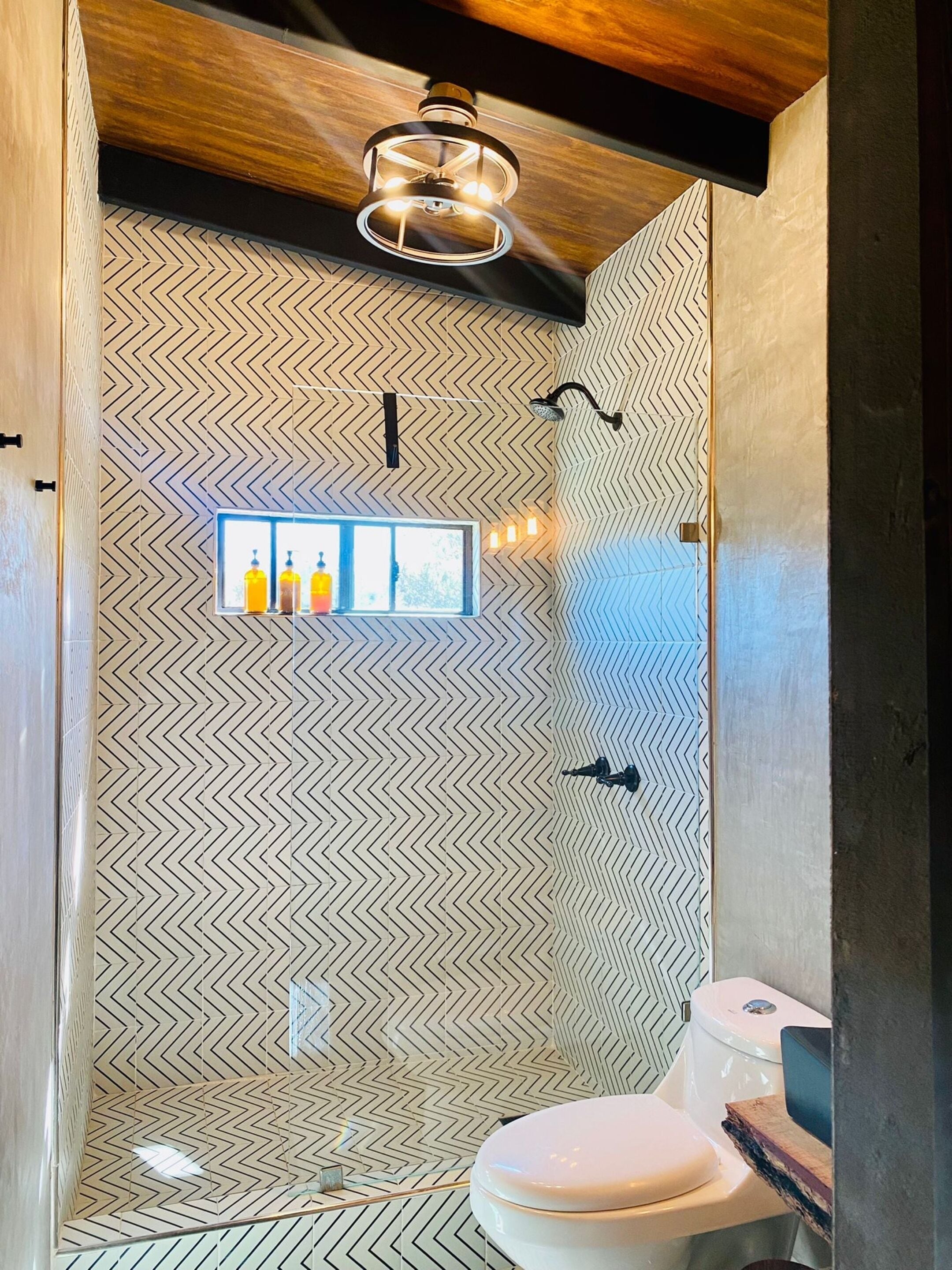 A bathroom with a chevron-patterned wall, a wooden ceiling, a toilet, and a shelf with bottles.