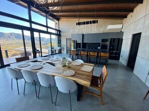 Una cocina moderna con una isla grande, un comedor con una larga mesa de madera y sillas blancas, y vistas a las montañas a través de amplias ventanas.